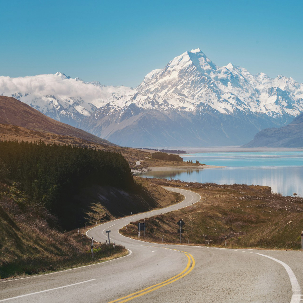 A two-lane road curving toward Aoraki Mount Cook past a glacial lake