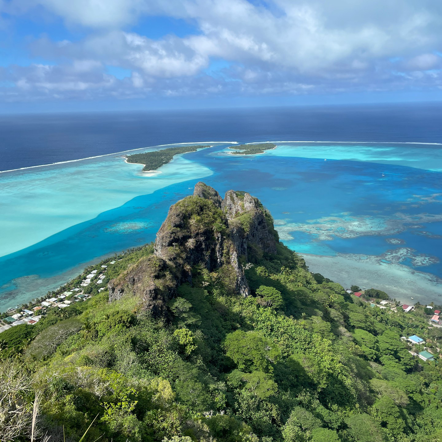 A green volcanic peak above a Polynesian lagoon, reef and motu beyond