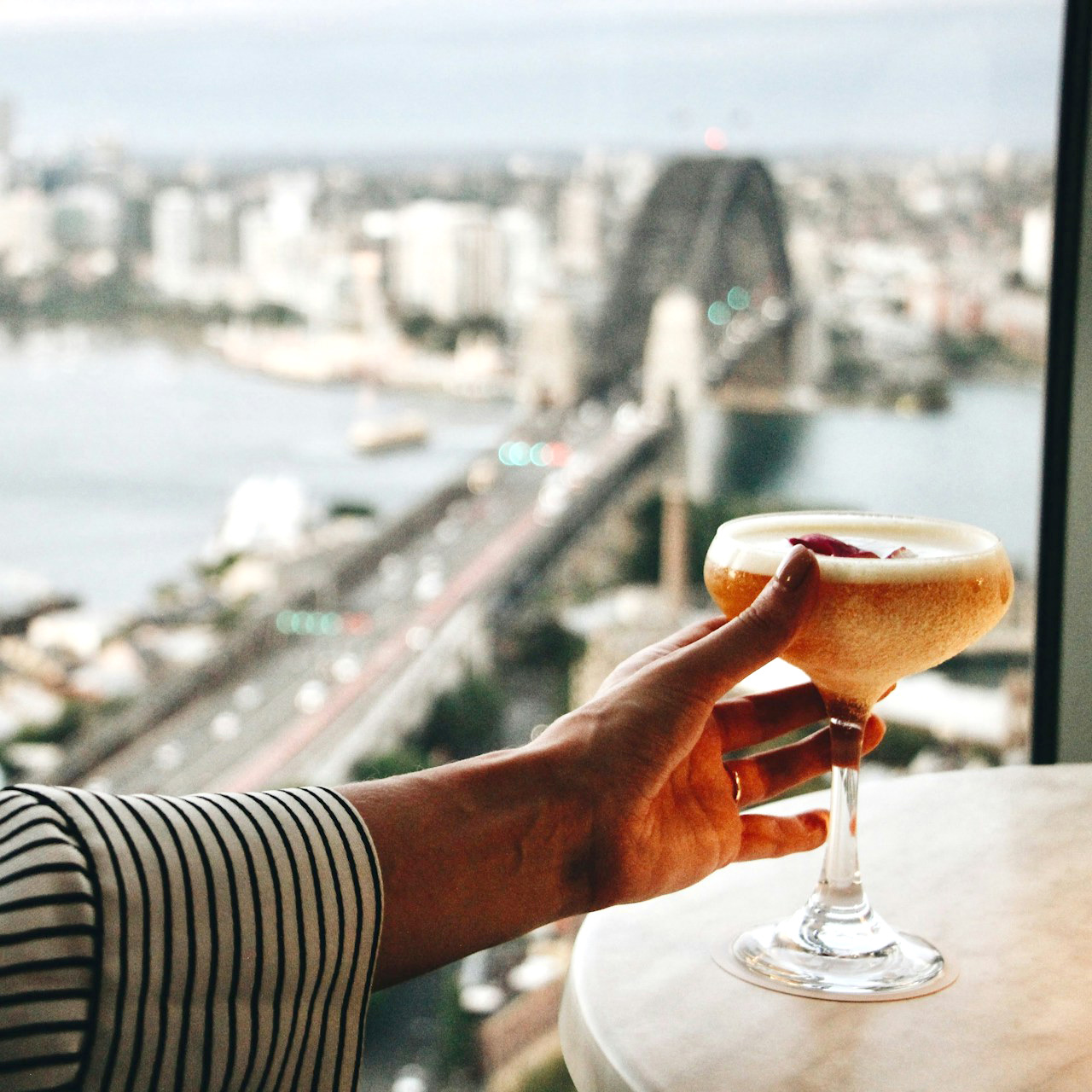 A drink held up against a blurred Sydney Harbour Bridge at dusk